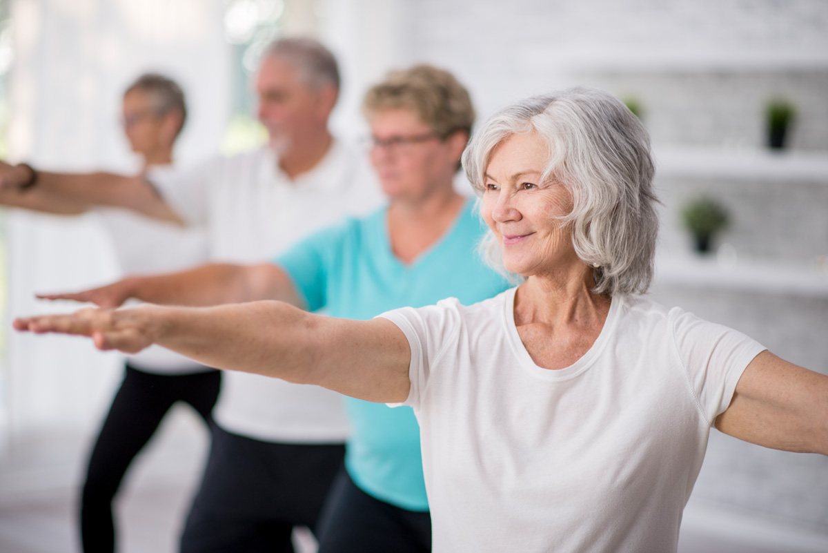 A multi-ethnic group of adult men and women are indoors in a fitness studio. They are wearing casual clothing while at a yoga class. A senior Caucasian woman is smiling while stretching out her arms.