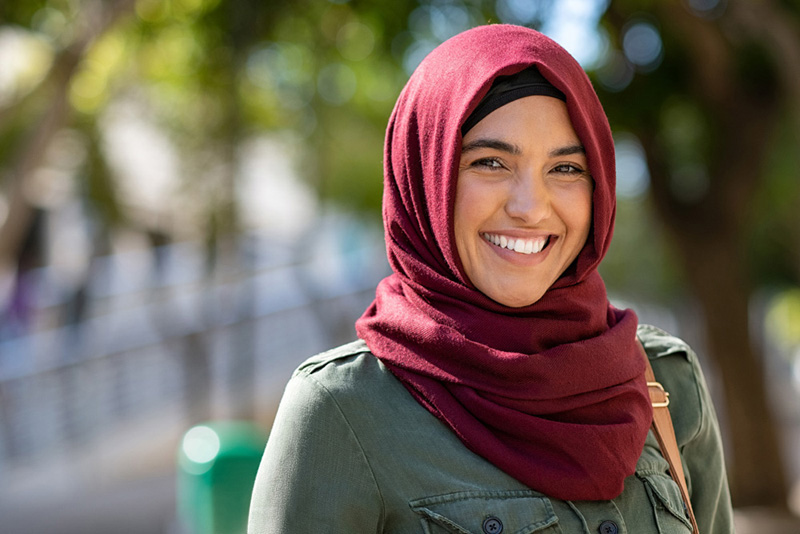 Portrait of young muslim woman wearing hijab head scarf in city while looking at camera. Closeup face of cheerful woman covered with headscarf smiling outdoor. Casual islamic girl at park.