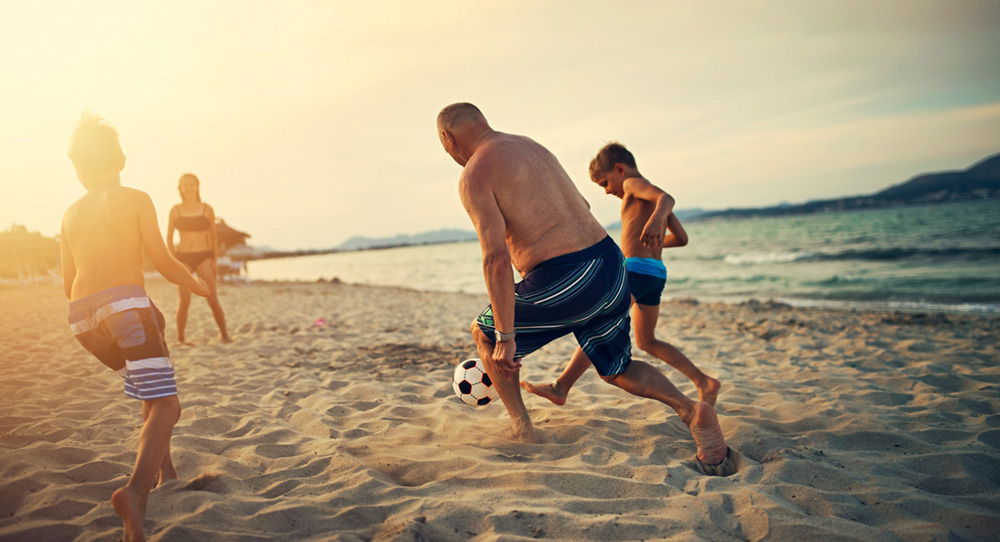 Three kids playing soccer with grandfather. The boys are aged 8 and girl is 13.  Family is enjoying sunny summer day on beach.
Nikon D850