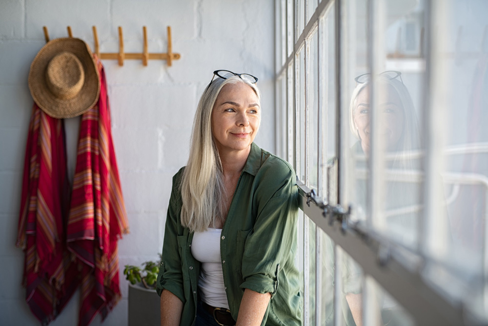 Smiling mature woman looking outside window. Thoughtful old woman looking away through window. Senior beautiful woman sitting at home with pensive expression.