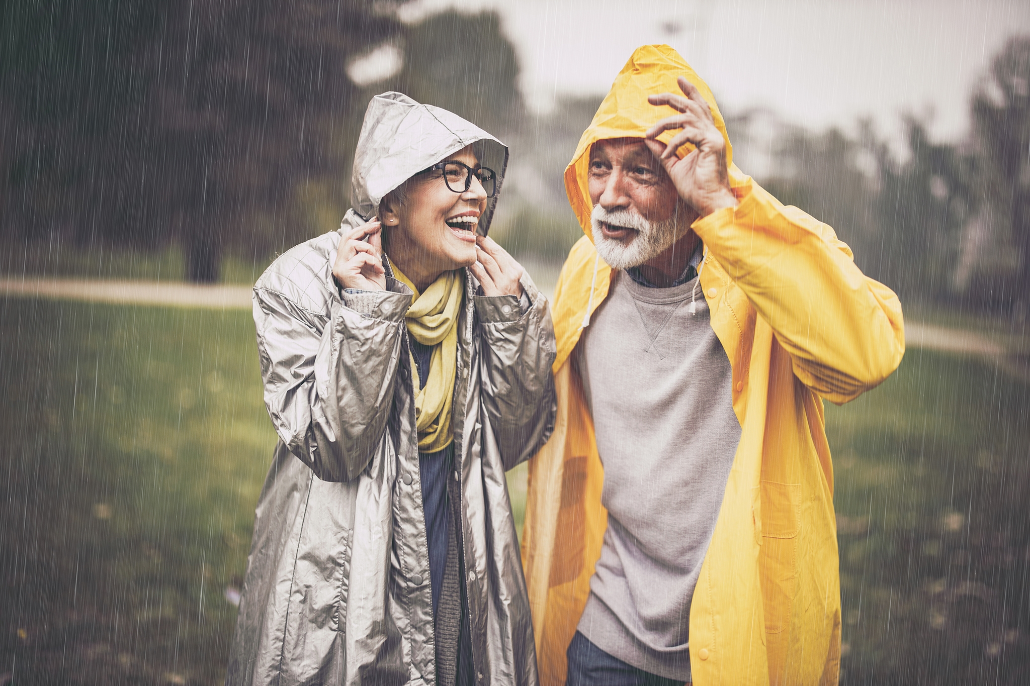 Happy senior couple in raincoats covering their heads during rainy day in nature.