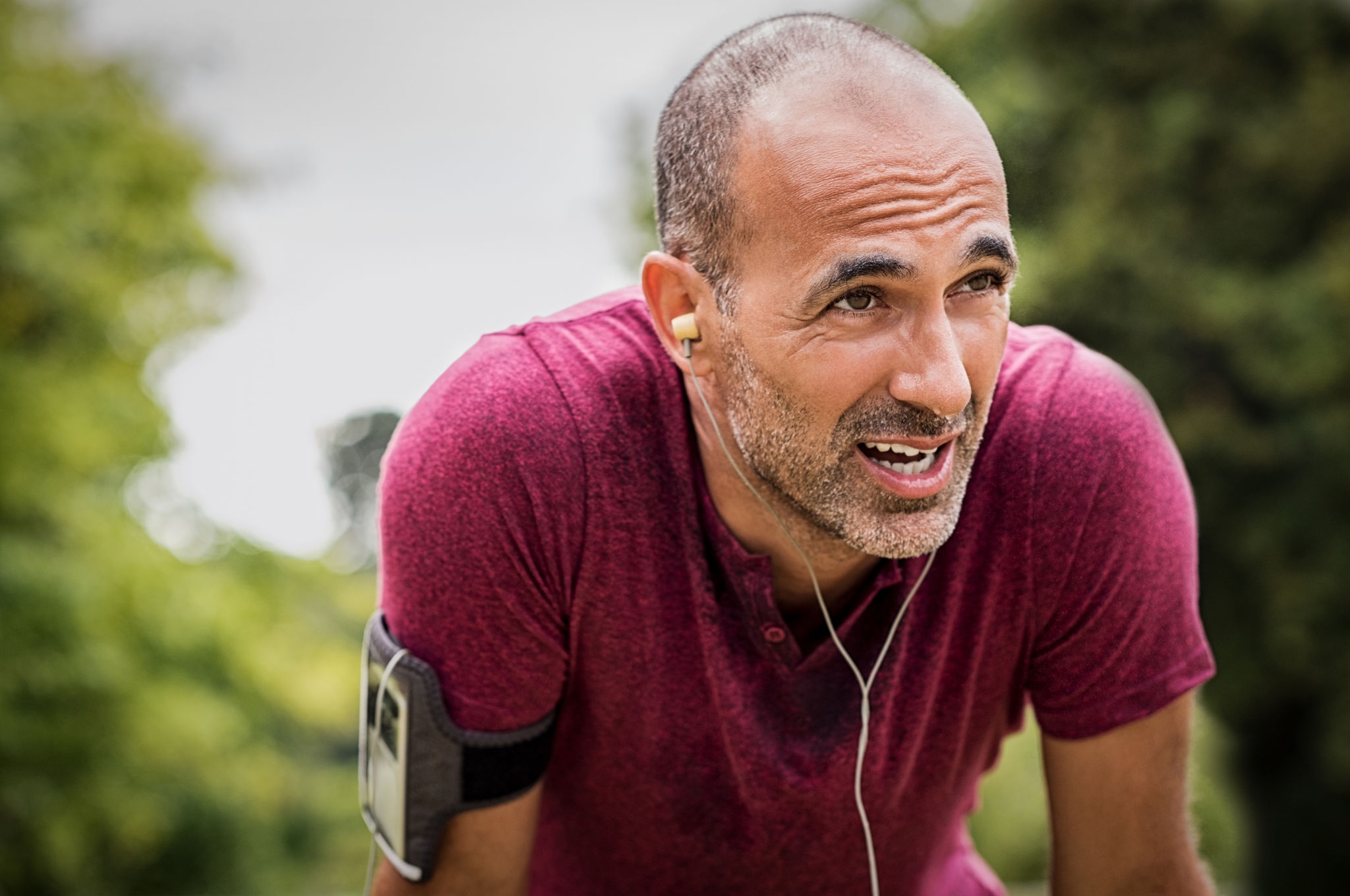 Portrait of athletic mature man after run. Handsome senior man resting after jog at the park on a sunny day. Sweaty multiethnic man listening to music while jogging.