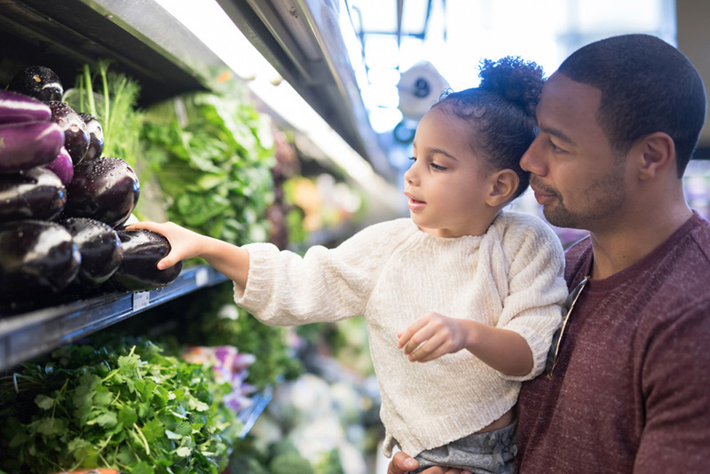 960043536 A pre-school age girl helps her dad pick out veggies in the produce section at the grocery store. He is holding her next to the produce and she is picking out eggplant.