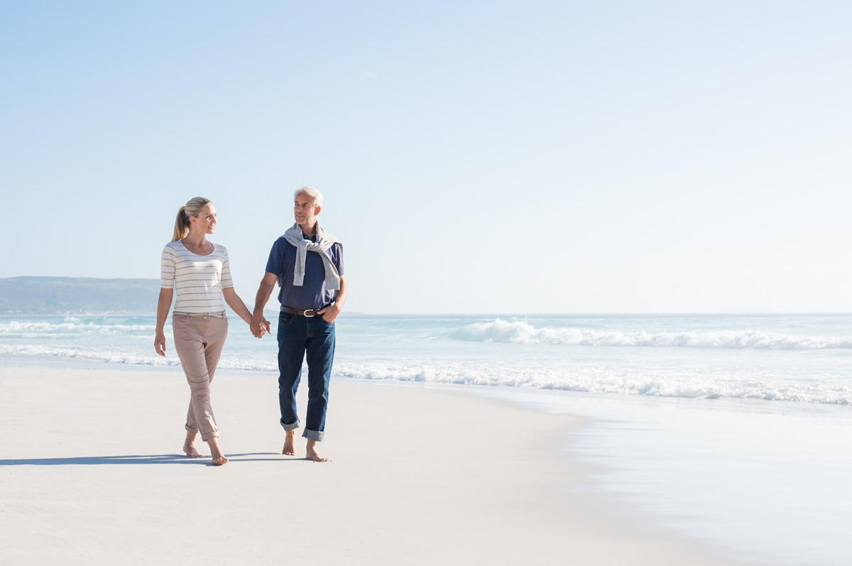 Senior couple holding hands at the beach on a bright sunny day. Mature couple in love holding hands and looking each other at the seaside. Smiling wife and happy husband walking barefoot on the white sand.