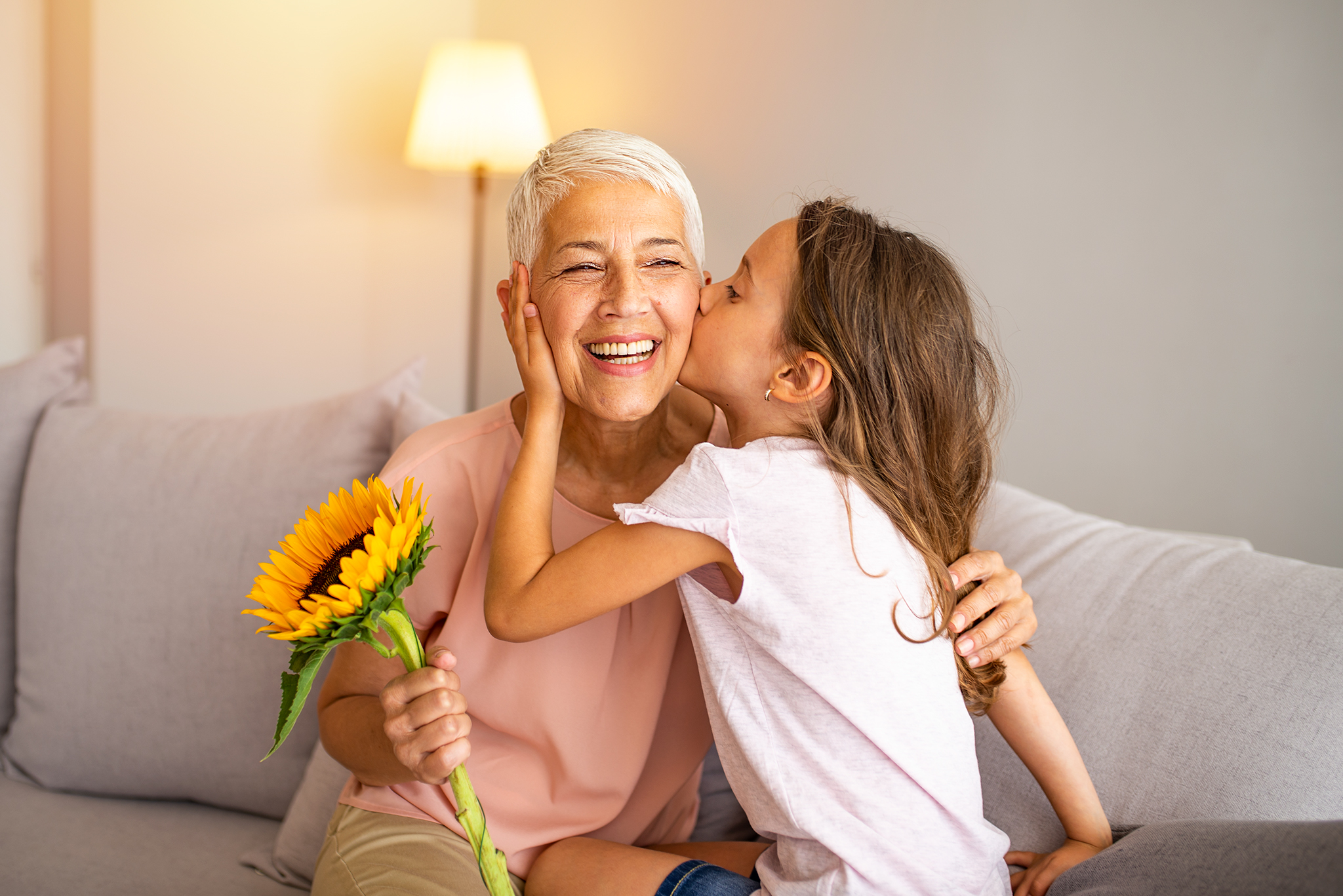 Little preschool granddaughter kissing happy older grandma on cheek giving violet flowers bouquet congratulating smiling senior grandmother with birthday, celebrating mothers day or 8 march concept