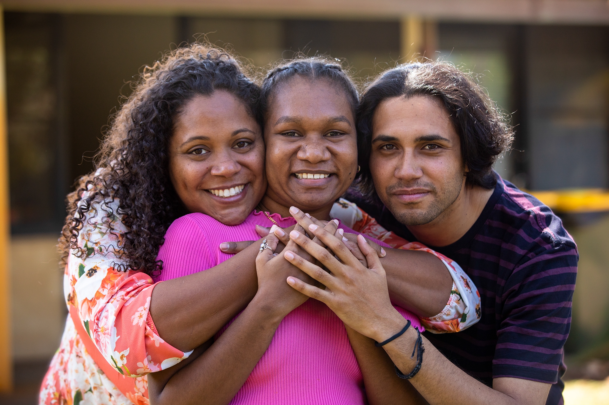 Three young aboriginal students outdoors with their arms around each other smiling at the camera.