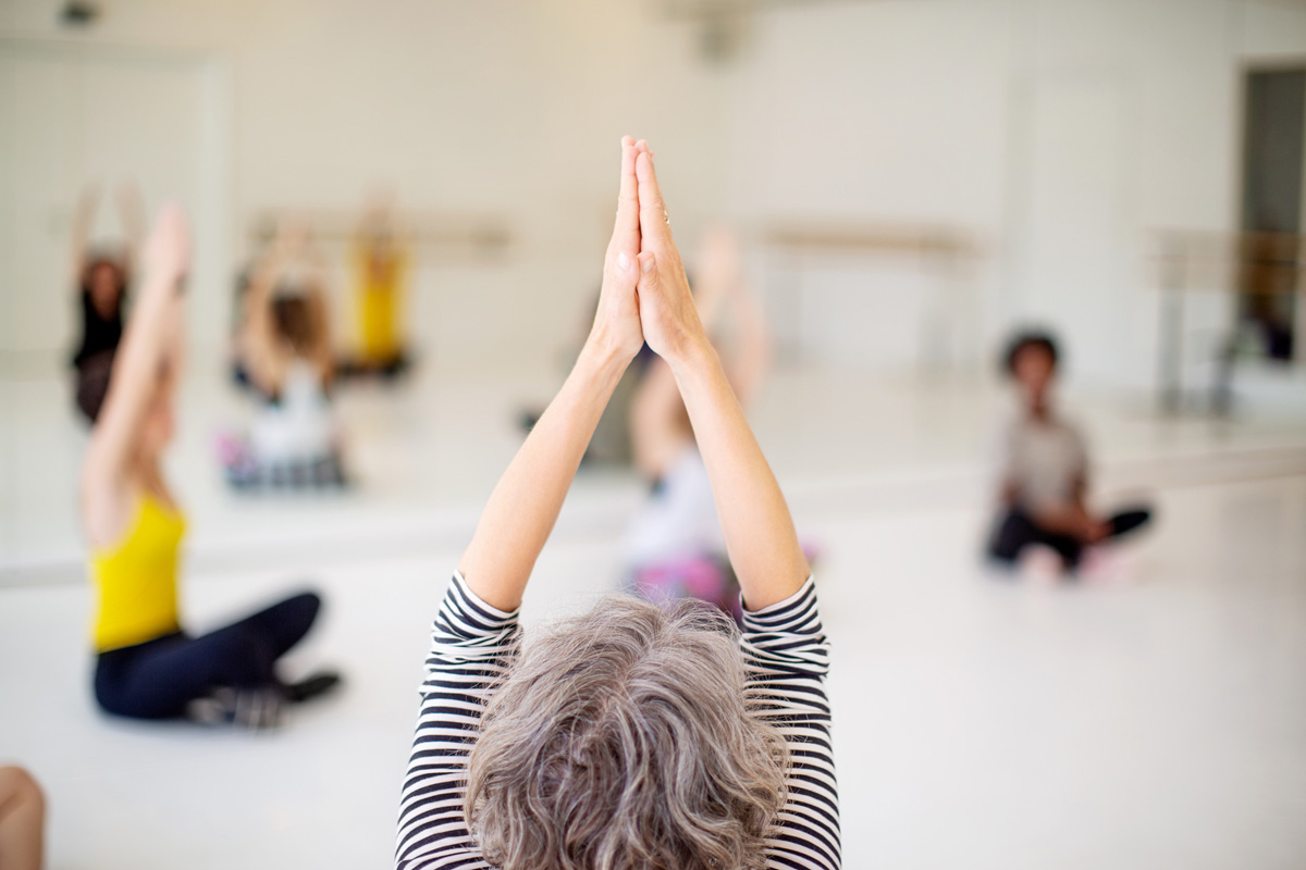 Shot of a mature woman sitting on floor with her hands joined above her head in fitness studio and other students exercising in background.