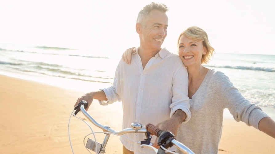 Mature couple cycling on the beach at sunset or sunrise. The ocean is in the background. They are happy and smiling. They are standing beside their bicycles. They are casually dressed. Could be a romantic retirement vacation.
