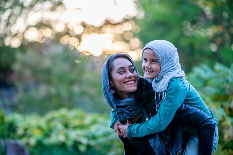 A Muslim woman and her daughter are outdoors in a public park on a sunny day. They are wearing casual clothes and head scarves. The mother is giving her daughter a piggyback ride.