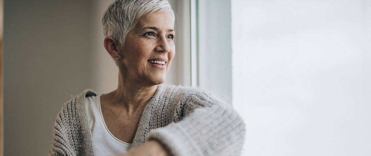 Happy senior woman day dreaming while looking through the window and enjoying in cup of coffee.