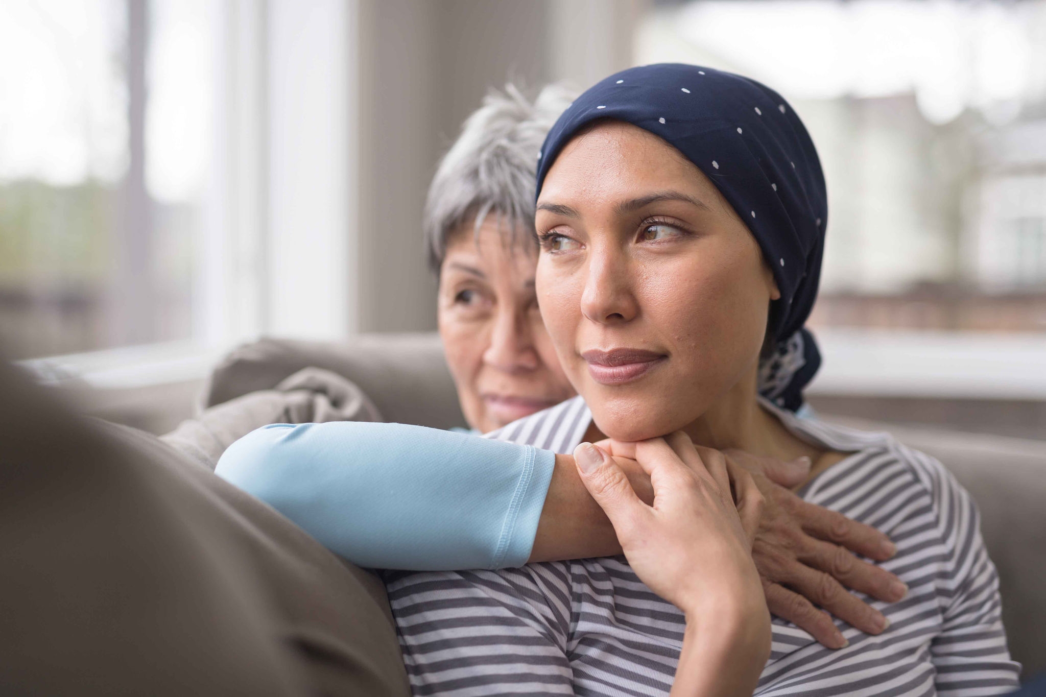 An ethnic woman wearing a headscarf and fighting cancer sits on the couch with her mother. She is in the foreground and her mom is behind her, with her arm wrapped around in an embrace, and they're both looking out the window in a quiet moment of contemplation.