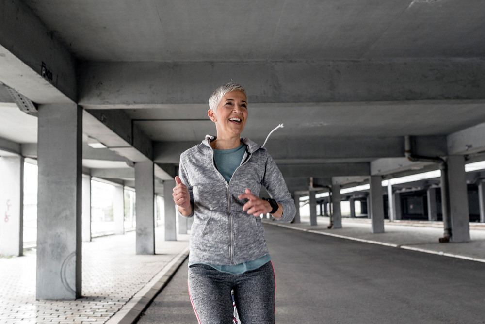 Photo of Smiling Mature, short hair, woman runner in the city in sports clothing looking away.  Active, happy gray hair, senior female jogging  for weight loss success during cloudy day. Starting day from morning jog, showing active retirement, exercising outdoor, positive, confident, energetic, focused, with toothy smile.