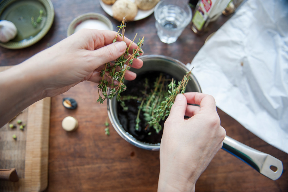 Woman adding fresh thyme leaves to a cooking pot filled with balsamic vinegar, to make a dressing. First person perspective.
