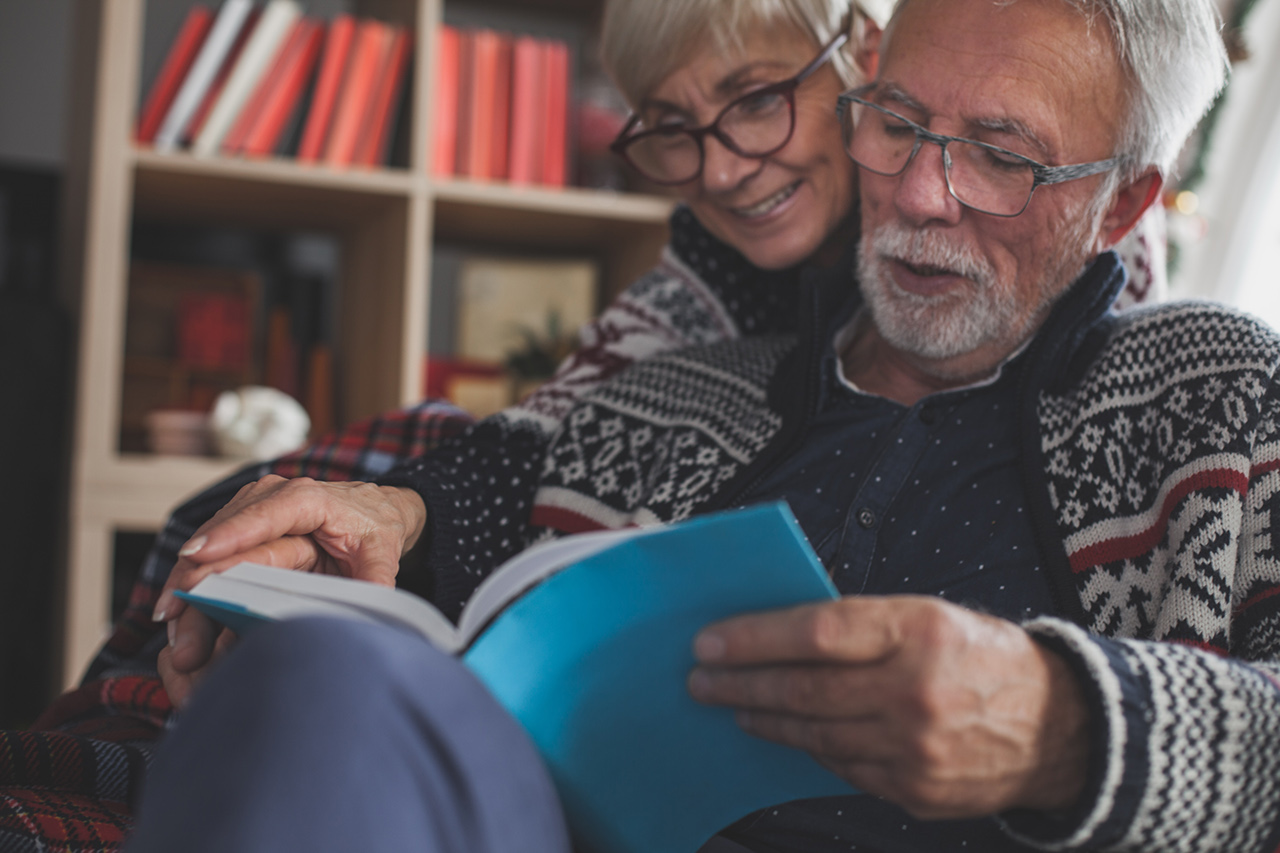 Senior married couple in cozy winters clothes relaxing and enjoying reading a book together.